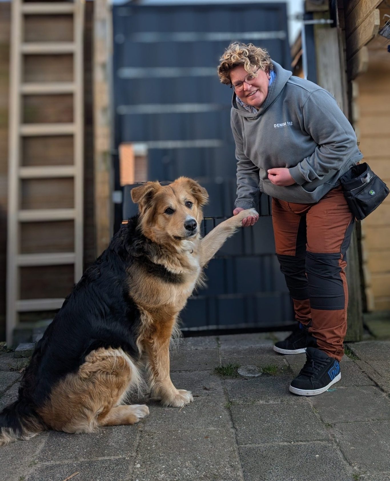 Man in een zwart shirt staat naast een blije zwarte hond in een groen veld.